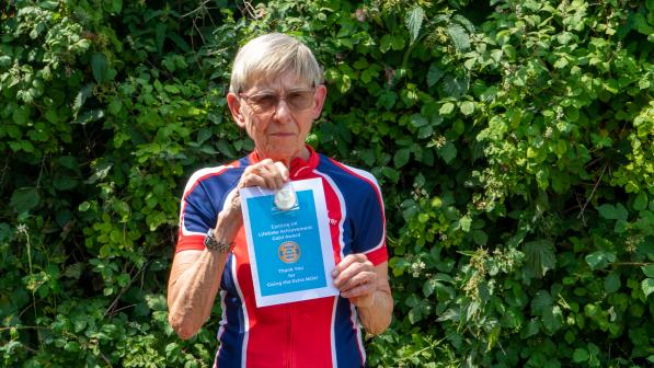 A woman wearing black cycling shorts and a red and blue horizontally striped cycling jersey is holding up a blue certificate.