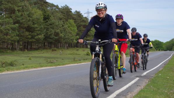 Four people are cycling along a quiet country road. All are wearing Cycling UK-branded T-shirts