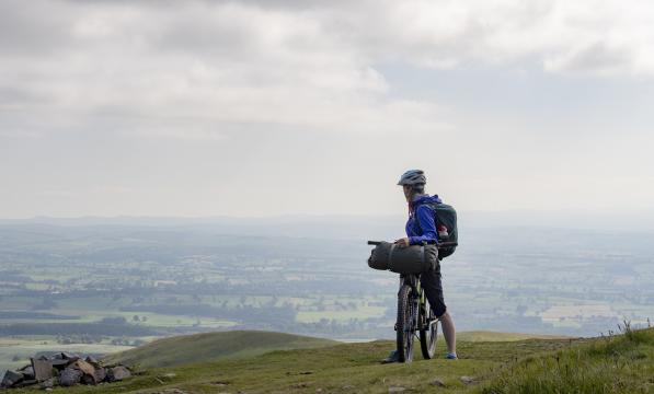A cyclist straddling her bike. She is looking back at rolling hills behind her. 