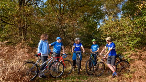Five cyclists standing on their bikes talking to each other while in a field with trees behind them