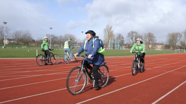 One white male and three white females are cycling along an athletics track. They have high-vis jackets on, and are wearing helmets and beaming smiles as they ride past the camera