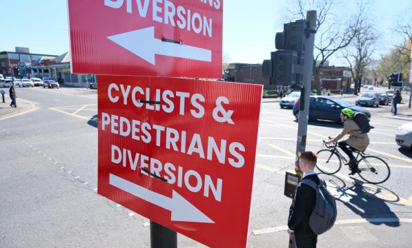 At a set of traffic lights, a red sign with bold white letters reads 'Cyclists and Pedestrians diversion' with an arrow pointing one way. The same sign appears facing the opposite way. In the background a pedestrian waits to cross a busy road