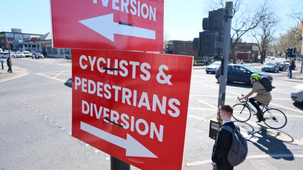 At a set of traffic lights, a red sign with bold white letters reads 'Cyclists and Pedestrians diversion' with an arrow pointing one way. The same sign appears facing the opposite way. In the background a pedestrian waits to cross a busy road