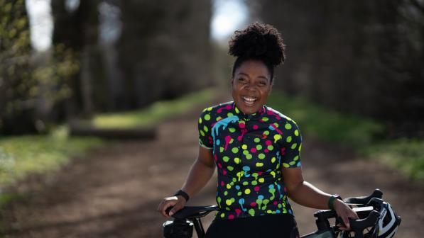 A woman is standing with her road bike. She is perched on the crossbar. She is wearing black cycling shorts and a black and multicoloured polka dot cycling jersey. She is smiling.
