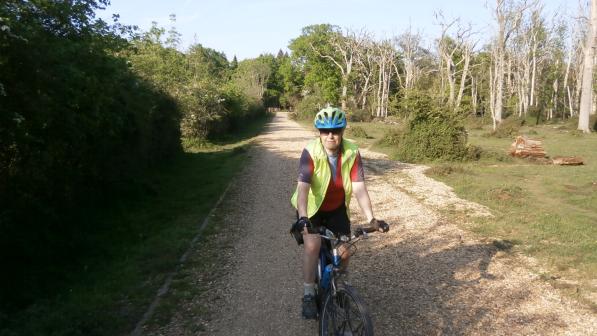 A woman is cycling towards the camera. She is wearing black shorts, an orange and grey jersey and yellow hi-vis jacket, along with a blue and green helmet and sunglasses. The bike is blue. She is cycling on a gravel path bordered by woodland on both sides