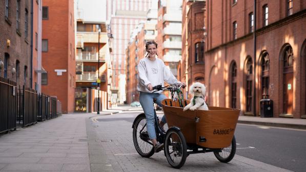 A woman is cycling on a cargo trike. She is wearing blue jeans and a white sweater. She has a NJ tube. A smally, fluffy white dog is in the cargo box.
