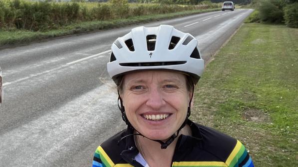 A woman is standing on a grass verge at the side of a road. She is wearing a white cycle helmet and a bright, stripey Stolen Goat cycling jersey. She is smiling at the camera.
