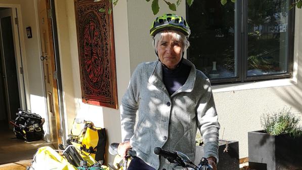 A woman is standing astride a grey hybrid bike with a packed trailer attached to it. She is wearing normal clothes: blue trousers and shoes and a grey fleece, as well as a cycle helmet.