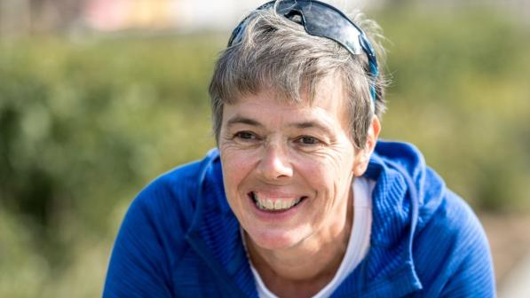 A woman is sitting on her bike with her forearms resting on the drop handlebars, her hands clasped in front of her. She is wearing a blue fleece and has sunglasses on her head. She has short hair. She is smiling.