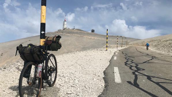 A loaded touring bike is leaning against a pole on a gravel path on a mountain