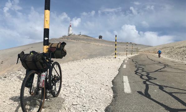 A loaded touring bike is leaning against a pole on a gravel path on a mountain
