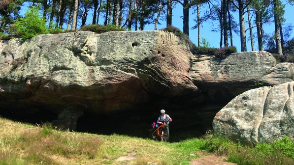 A man is wheeling an orange mountain bike away from a cave. He is wearing cycling kit and a helmet