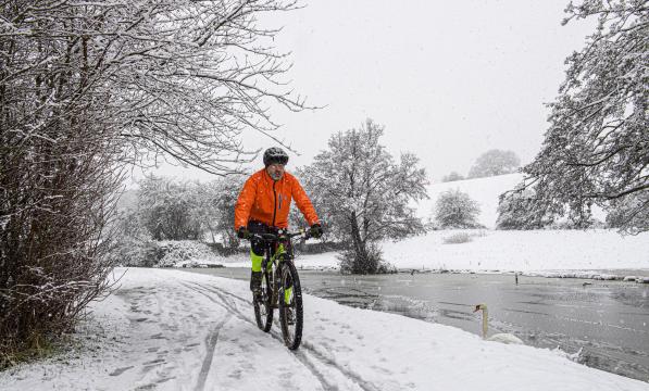 A man is cycling along a snowy path next to a frozen river. It’s a very wintry scene, it’s snowing. He is wearing a bright orange winter jacket and black winter cycling leggings with bright yellow sections. There’s a single swan on the river