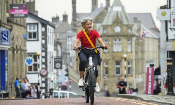 A woman is riding along a town centre road on an e-cycle. She is wearing a red T-shirt and black three-quarter length trousers with a messenger bag across her body. There are shops and businesses in the background