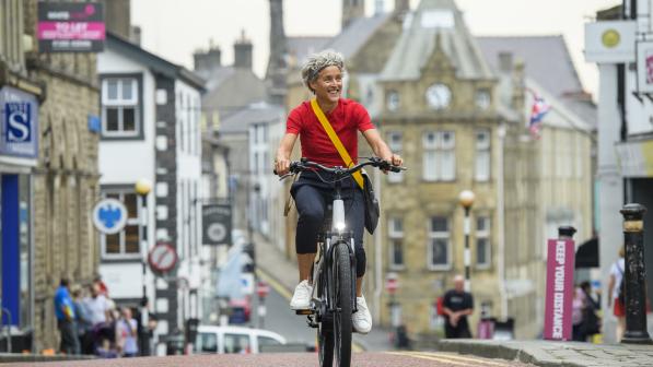 A woman is riding along a town centre road on an e-cycle. She is wearing a red T-shirt and black three-quarter length trousers with a messenger bag across her body. There are shops and businesses in the background