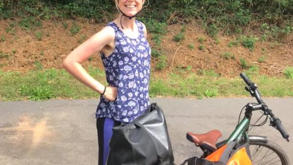 A woman is standing next to a touring bike. She is wearing a blue strappy top with white flower pattern and blue and black leggings, a turquoise cycling helmet and sunglasses. The bike is green and it has two big panniers on the rear rack