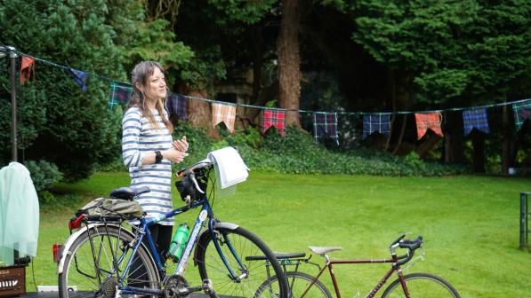 A woman is standing on a makeshift stage. She has a Dawes touring bike with her; an open notebook is balanced on the bike’s handlebars. She is wearing a blue and white striped top, black three-quarter-length trousers and cycling shoes. She is smiling.