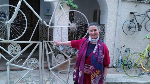 A woman is standing at the gates of a hotel in Spain. The building is covered with old bicycles and even the gates are made from bike frames. She is wearing black trousers, a bright cerise cardigan and blue top, as well as a brightly patterned scarf