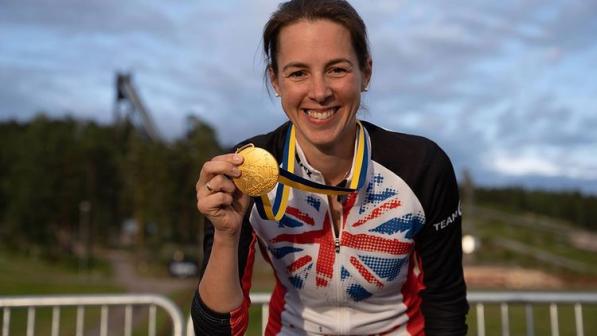 A woman in a GB cycling jersey is holding up a gold medal and smiling to the camera 