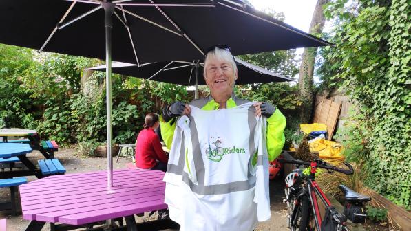 A woman is standing in a pub or restaurant garden. She is wearing cycling clothing and gloves. She is holding up a JoyRiders jacket