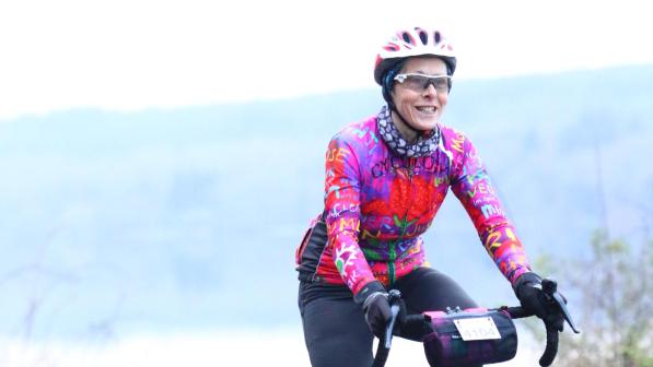 A woman is cycling along the road. She is on a road bike which has a number attached to a handlebar bag; she is taking part in an organised cycling event. She is wearing black cycling leggings and a very bright multicoloured jersey