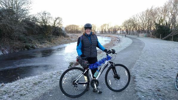 A woman is standing with a light grey Trek mountain bike. It’s winter. She is wearing a warm winter jacket and trousers. Her bike is decorated with blue tinsel. There is frost on the pavement, grass and trees and the stream behind her has ice on it