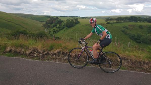 A woman is cycling on an Orbea road bike. She is wearing a green and white cycling jersey, black shorts, a red and white helmet and black and yellow fingerless gloves. She is smiling at the camera. In the background is some pretty impressive countryside