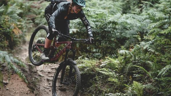A woman is cycling downhill through a forest. She is riding a Scott mountain bike. She is wearing black three-quarter-length cycling leggings and a black jacket, as well as a green helmet. Credit: Roo Fowler