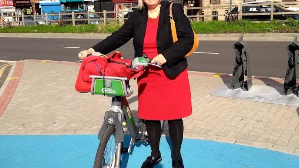 A woman is standing with a hire bike on some of Birmingham’s cycle infrastructure, the blue routes. She is wearing a red dress, black cardigan and black tights. She has a yellow handbag over her shoulder and a red bag in the bike’s basket