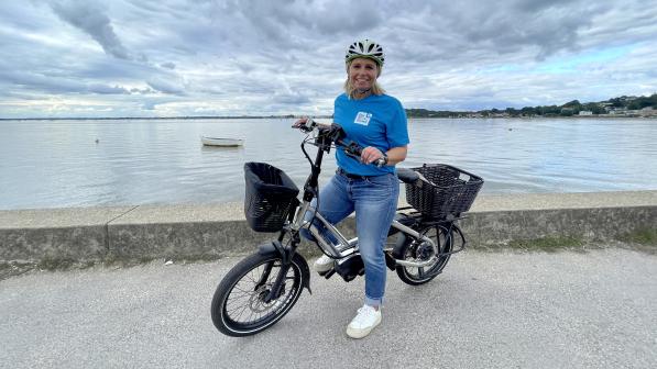 Lucie on a silver e-cycle with front and back baskets, standing in front of Poole harbour. She is wearing blue jeans and a blue BH Active Travel T-shirt