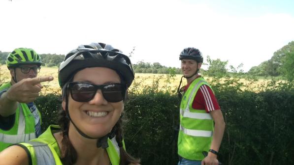 A selfie of three cyclists next to a hedge. The photo has been taken by a woman and behind her are two men, one to the left and one to the right. They are all wearing hi-vis jackets and cycling helmets