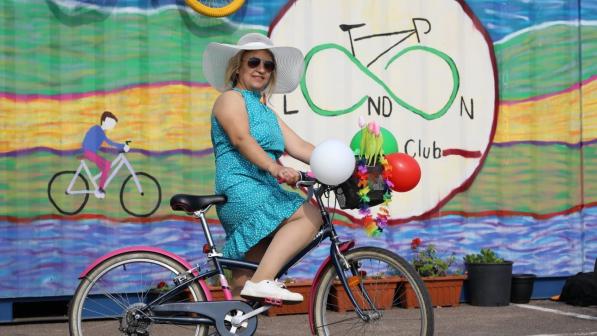 A smiling woman with long blonde hair and sunglasses is standing astride a black and pink bike. She is wearing a blue dress and white sun hat. The bike’s basket is decorated with balloons and plastic flowers