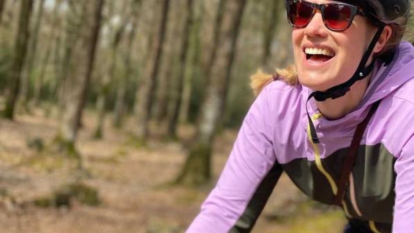 A woman in a lilac and forest green cycling jacket and silver helmet is cycling off road through a forest. It’s autumn and the trees are bare, there are leaves all over the forest floor
