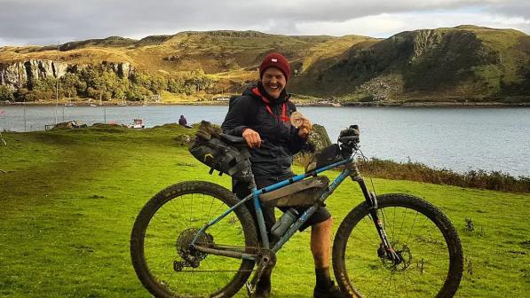 A woman is standing with a very muddy bike and holding a medal, having just completed an off-road triathlon on the Isle of Kerrera. She is smiling broadly