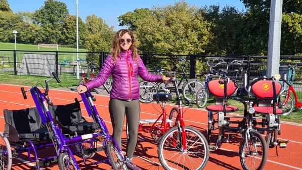 A woman is standing on a racetrack. She is wearing black leggings and a violet padded jacket, along with sunglasses. She has long, wavy, dark blonde hair. She is surrounded by various non-standard cycles, showing the different options for different needs