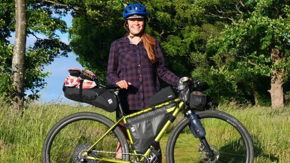 A woman is standing with a metallic green bike that is fully loaded with bags for bikepacking. She is wearing a blue cycling helmet and a check shirt. She is smiling at the camera