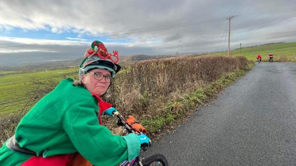 A woman is riding a bike while wearing a Christmas elf outfit. She is looking back at the camera and pulling a funny face