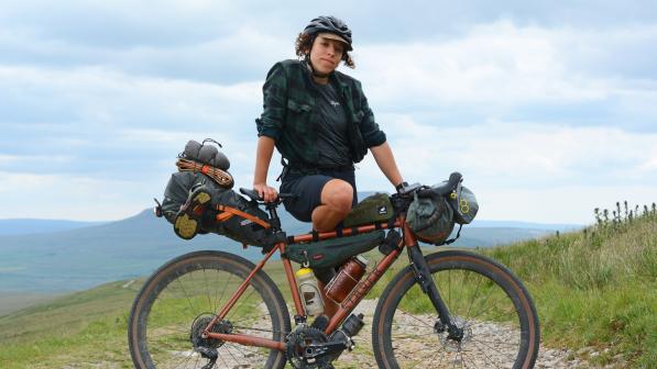 A woman is posing with a heavily laden bikepacking bike. She is wearing black shorts, a black Rapha T-shirt and check shirt, and a black cycling helmet with a cap underneath. She is in a remote location with mountains in the background