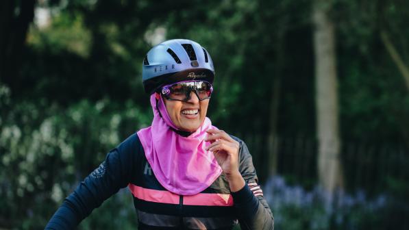 A woman in cycling gear is smiling at the camera. She is wearing a blue cycling helmet with a Rapha cap underneath. She has on cycling sunglasses, a stripey Rapha cycling jersey and a bright pink headscarf