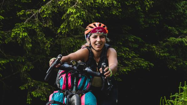 A woman is pushing her bike uphill. Only the bike’s handlebars and top of the front wheel are in shot, along with packed bikepacking bags. She is wearing an orange helmet and a Rapha cap