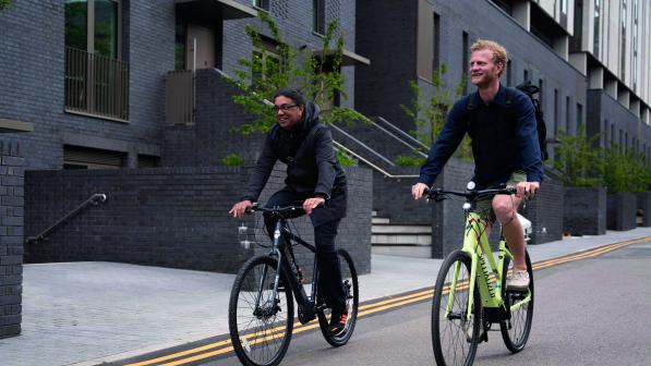 Two men are riding e-cycles side by side along an urban street. They are wearing normal clothes with no cycling helmet.