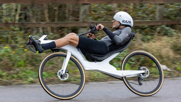 A man is riding a white recumbent cycle around a track. He's wearing a white cycle helmet and black and grey racing kit