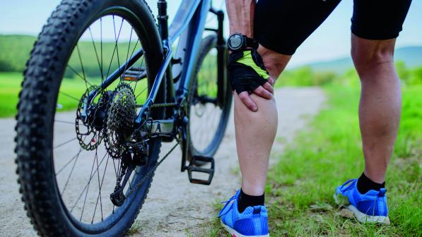 Seen from behind, a man is standing by the side of a road with a mountain bike in front of him. He's bending over and holding the back of his knee as if it hurts