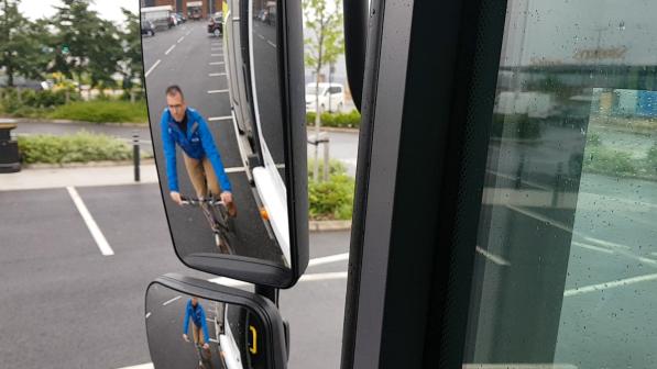 A close-up of a left-hand-side wing mirror on a lorry showing a man on a bike in the background