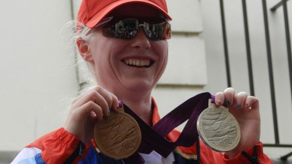 Paralympic cyclist Aileen McGlynn holding her silver and bronze medals won at the 2012 Summer Paralympic Games. She is wearing Team GB colours, a red peaked cap and cycling sunglasses.  Photo: The Rambling Man and Kim Ratcliffe of Think Equestrian under the Creative Commons Attribution-Share Alike 3.0 Unported license. https://creativecommons.org/licenses/by-sa/3.0/deed.en