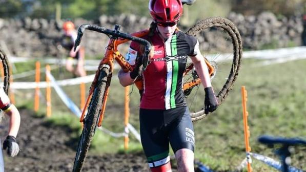 Aisling Charlesworth carries her cyclocross bike across a muddy section of a race. She is wearing red, white, green and black Fibrax Wrexham Roads Club kit, a helmet and sunglasses. She is covered in mud!