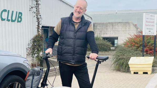 A man in black trousers, bodywarmer and long-sleeved T-shirt is holding a white Brompton bike