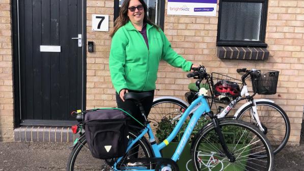 Alison Holland is standing outside a brick building. She is holding a blue hybrid bike with a front basket and rear panniers. She is wearing a green fleece and black trousers. She has long brown hair and sunglasses. Behind her is another bike, a white hybrid.