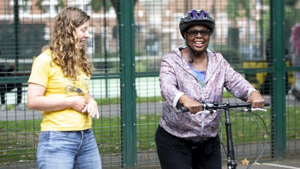 Alison Litherland, a white woman with curly hair wearing a yellow t-shirt, stands smiling next to a Black woman who is wearing a helmet and holding a bicycle. They are outdoors in a fenced area, engaged in a friendly conversation.