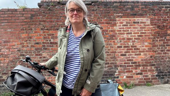 Alison Stenning is standing in front of a brick wall, holding her black hybrid bike behind her. It has a front basket, rear pannier and frame bag. She is wearing a green jacket, stripey top and black jeans.